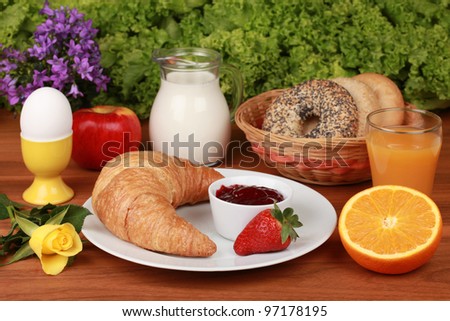 French breakfast with a croissant and strawberry jam. Served with milk, orange juice, egg, apple, bagels and flowers.