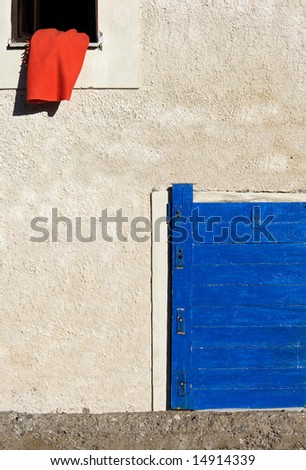 Image shows a red blanket on the window of a Greek village house. A blue door appears in the lower right corner