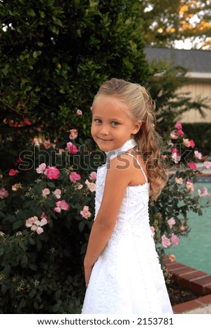 A pretty little girl dressed in a long white dress.  She has blonde hair and blue eyes and is standing by a bed of pink roses. Her Hair has long flowing curls.