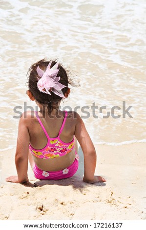 Little girl with big pink bow in her brunette hair sitting in the sand on the beach watching the waves.