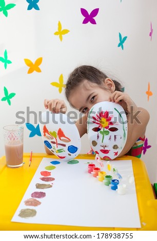 A girl holding two painted masks. The child in bright clothes in playroom