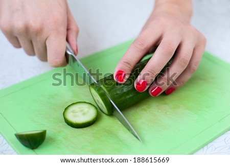 Closeup female hands cutting cucumber on cutting board, red manicure, strong contrast