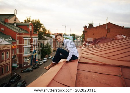Young bold girl woman in hipster clothes, sit on edge of roof, dressed like a boy man in shirt, bow-tie, suspenders and pants trousers on background of city, road and jam