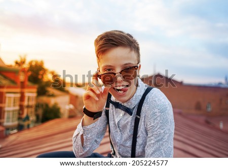Young bold girl woman in hipster clothes, sitting on roof showing off tongue looking above sunglasses