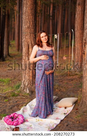 Beautiful happy pregnant woman outdoors in pine coniferous forest wearing romantic dress and holding her belly, scene is decorated in rustic style
