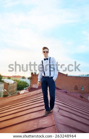 Young bold guy boy girl woman in hipster clothes on edge of roof, dressed like a boy man in shirt, bow-tie, suspenders and pants trousers on background of city