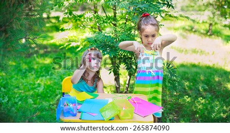 Girls, sisters, kids, friends cutting multicolored paper outdoors, classroom playing in the garden. Focus on elder girl\'s face, and on younger girl\'s hand with scissors