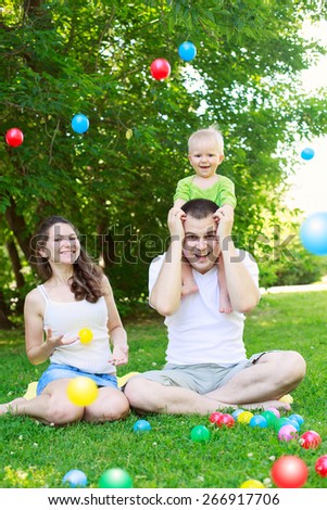 Happy family outdoors  mum and dad hold baby, playing with colorful balls, sitting on grass