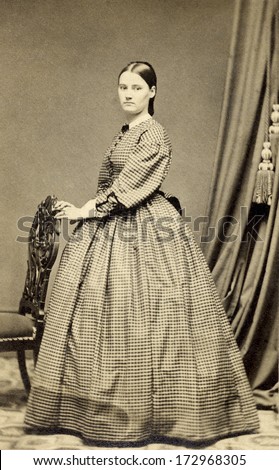 USA - CONNECTICUT - CIRCA 1864 - A vintage Cartes de visite photo of young pioneer woman standing with hand on chair and dressed in hoop skirt dress. Photo from the Civil War Victorian era. CIRCA 1864