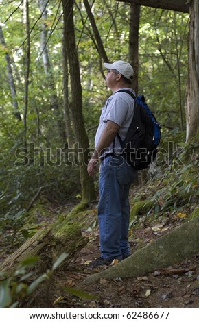 A middle aged man hiking out in the woods with a back pack and base ball hat wearing blue jeans and tennis shoes. He is looking at the forest around him while taking a break.