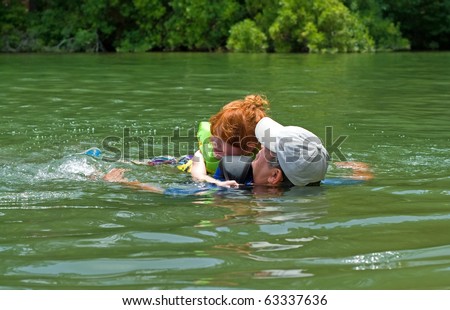 A mature man with his granddaughter swimming in a lake. Both the grandfather and child have on life vests for safety.
