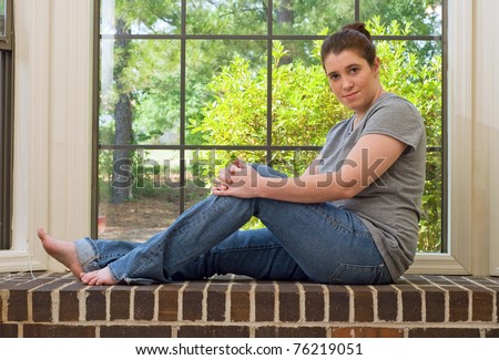 A young woman sitting on a brick ledge in front of a window wearing a gray shirt and blue jeans.