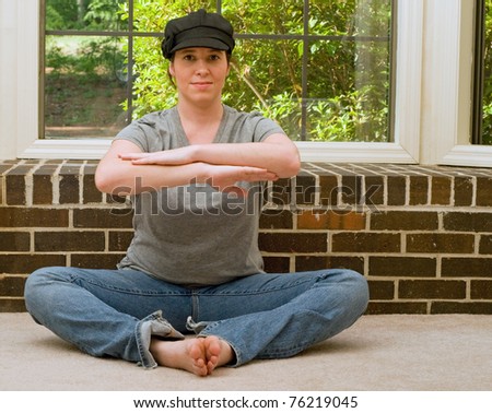A young woman sitting and relaxing while doing Yoga in front of a brick ledge with a window behind it. She is wearing a black hat and a gray shirt with blue jeans