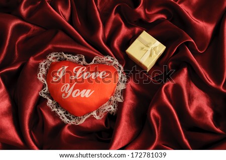 Heart with I love you written on it and a small jewelry gift box on a red satin sheet