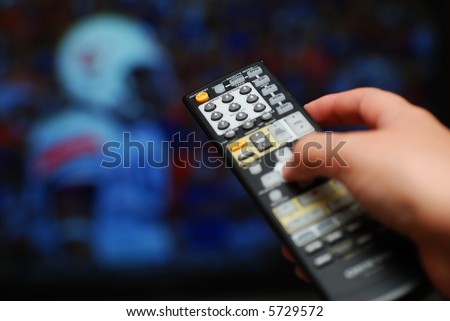 A hand holding a TV remote control and pointing it toward a television showing a football game. Selective focus. TV image is out of focus.