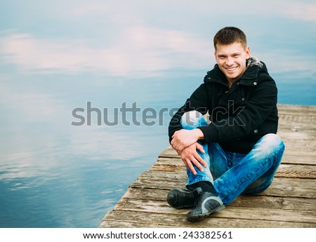 Young handsome man sitting on wooden pier in autumn day, relaxing,  thinking, smiling. Casual style - jeans, jacket