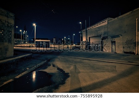 Dark factory warehouse alley with railroad tracks and rain puddle at night.