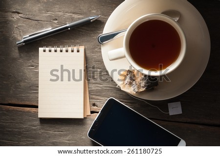 Small notepad with a cup of tea, pen, pencil and cellphne on rustic wood background with low key scene