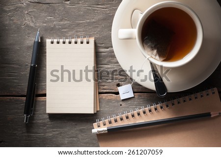 Small notepad with a cup of tea, pen and pencil on rustic wood background with low key scene