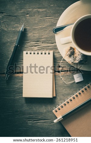 Small notepad with a cup of tea, pen and pencil on rustic wood background with film filter effect