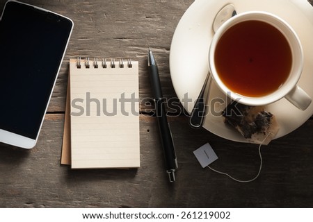 Small notepad with a cup of tea, pen, pencil and cellphne on rustic wood background with low key scene
