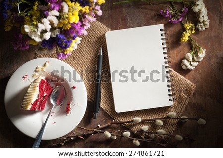 Small notepad with pen, pencil and bits of red velvet cake on rustic wood background with low key scene.
