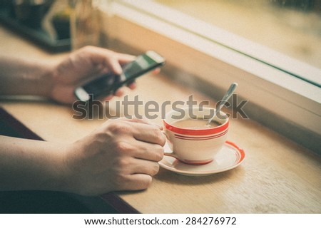 Male hand holding a cup of coffee on wood bar with another hand using smartphone in blurry background with film filter effect