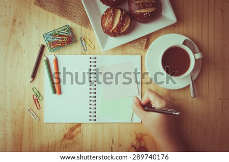 Notebook and sticky paper, paper clip, color pencil, and pen on wood table in vintage style with female right hand wrting on notepad with film filter effect