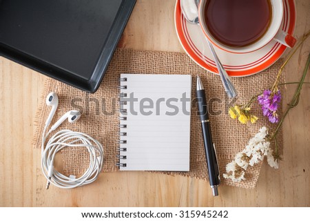 Opened notepad with pen, empty black ceramic dish, small earphone, and a cup of hot tea on wood table in cafe with morning scene