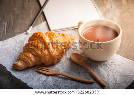 Butter croissant with a cup of hot chocolate and small notebook on wood table with morning scene