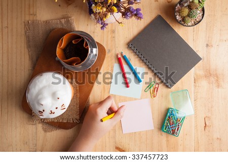 Woman left hand writing on sticky paper with blank space for text or message on wood table with notebook, paper clip, color pencil, and pen in cafe