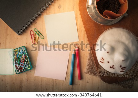 Sticky paper with blank space for text or message, notebook, paper clip, color pencil, and pen on wood table with iced thai tea cup and coffee drip cup beside at cafe in morning time