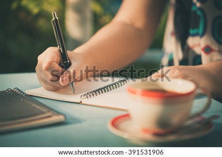Woman hand writing journal on small notebook at outdoor area in cafe with morning scene and vintage filer effect