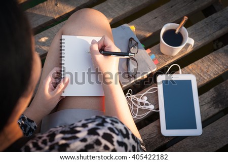 Young hipster woman writing journal on small notebook while sitting on wood bridge in morning time on weekend with high contrast sun lighting