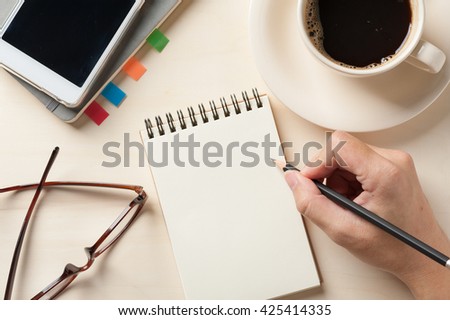 Young man right hand writing on blank notebook on wood table with coffee cup, smartphone, and glasses beside in morning time