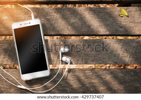 Smartphone with blank area on touch screen and in ear headphone on rustic wood table at outdoor area in morning time with high contrast natural lighting