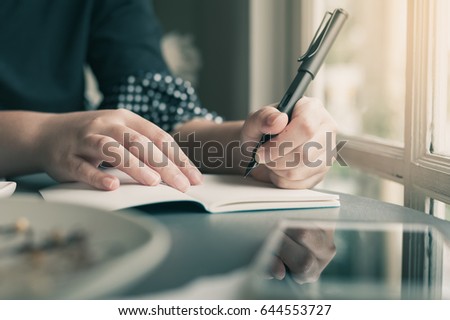 Left-handed woman hand holding pen while writing on small notebook beside window. Freelance journalist working at home concept with vintage filter effect