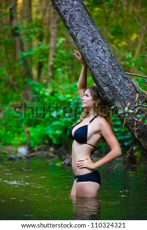 young woman in bathing suit with big breasts is in the water in woods near tree on a green background