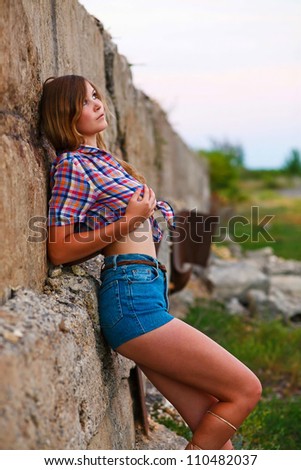 blonde young woman in cowboy shirt and shorts, his hands on his chest to wall, looking into distance