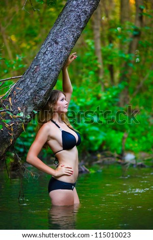 young nude woman in bathing suit with big breasts is in the water in woods near tree on a green background
