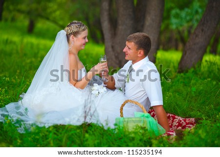 bride and groom at wedding in green forest sitting on picnic, drink wine from wine glasses at wedding