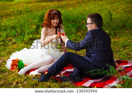 Redhead bride and groom at wedding in green field sitting on picnic, drink wine from wine glasses at wedding