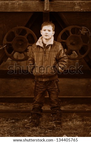 Retro black and white photo of sepia boy blonde tramp in a brown jacket, crumpled jeans on the street near the railway wagons