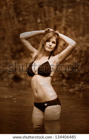 Retro black and white photo of sepia Blonde young woman with big breasts in a black bikini standing on a river in the woods with his hands behind his head