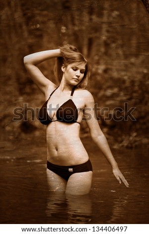 Retro black and white photo of sepia Blonde young woman with big breasts in a black bikini standing on river in woods with his hands behind his head