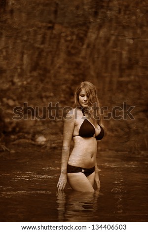 Retro black and white photo of sepia Blonde nude young woman big breasts in a black bikini standing on a river in the woods with his hands behind his head