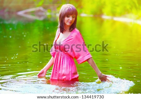 sunlight brunette woman red dress is wet to waist in water touches hand of river
