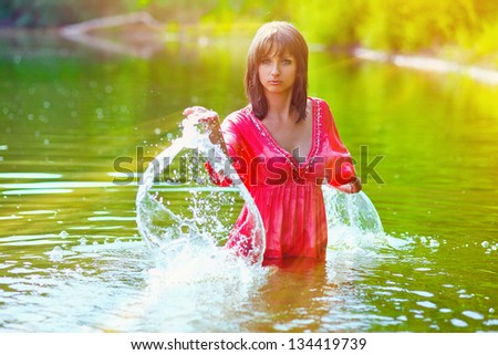 sunlight brunette woman red dress is wet to waist in splash water touches hand of river