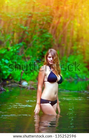 sunlight Blonde nude young woman big breasts in a black bikini standing on a river in the woods with his hands behind his head