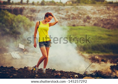 homeless young beautiful woman in a yellow tank top and denim shorts standing on a landfill in the smoke, pensive and serious Grusno